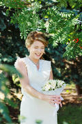 Девушка в белом платье в тени деревьев с маленьким белым букетом на свадьбе