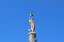 Фигурку журавлика на пне дерева напротив голубого неба.